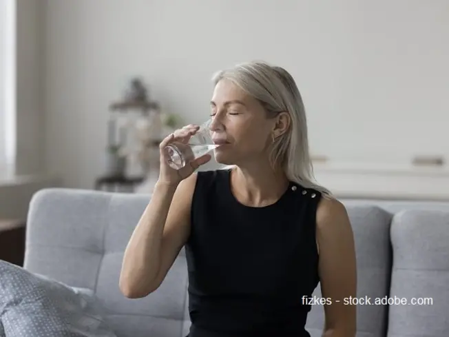 Bild einer Frau mit grauen Haaren, die auf der Couch sitzt und ein Glas Wasser trinkt.