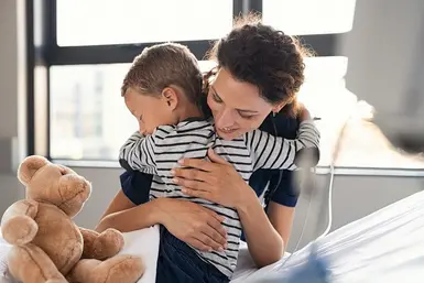 Kleiner Junge in gestreiftem Pullover auf Krankenhausbett umarmt glücklich Krankenschwester mit dunklen Haaren. Neben ihm liegt ein Teddybär.