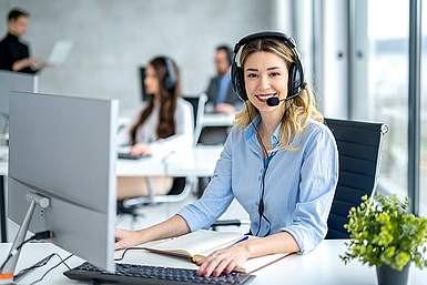 Eine junge Frau mit blonden mittellangen Haaren sitzt im Büro an ihrem Schreibtisch. Sie trägt ein Headset und vor ihr steht ein Bildschirm.