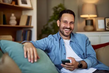 Erleichterter und glücklicher Mann sitzt auf der Couch mit seinem Handy in der Hand weil er soeben die Krankentagegeldversicherung des Münchener Vereins abgeschlossen hat.