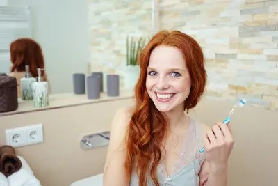 Junge Frau mit rotem Haar hält eine Zahnbürste und lacht in die Kamera.
