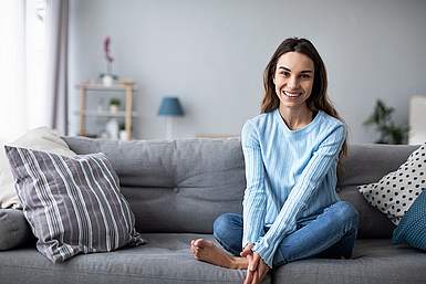 Junge Frau mit braunen Haaren und blauem Pullover sitzt auf der Couch und lacht in die Kamera.