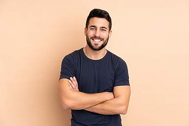 Junger Mann mit Bart und dunklem T-Shirt vor beigem Hintergrund lacht in die Kamera.