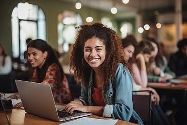 Eine junge Frau mit Locken sitzt lachend und mit verschränkten Armen an einem Tisch vor ihrem Laptop.