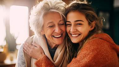 Junge Frau mit braunen Haaren und orangenem Pullover umarmt glücklich ältere Frau mit grauen Haaren.