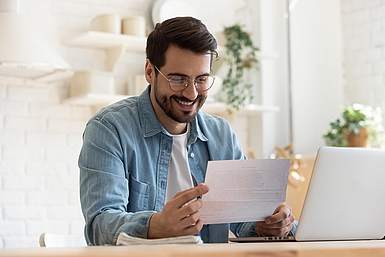 Ein junger Mann mit kurzen braunen Haaren und Brille sitzt am Tisch und hält einen Brief in der Hand, welchen er erfreut durchliest.