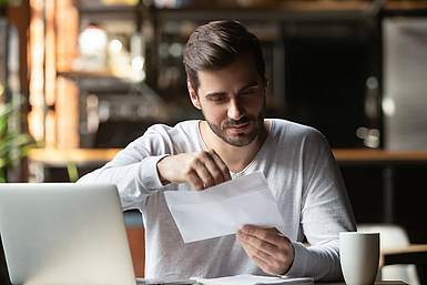 Ein junger Mann sitzt an seinem Schreibtisch. Vor ihm steht sein Laptop und in der Hand hält er eine Brief, den er gerade in ein Kuvert steckt.