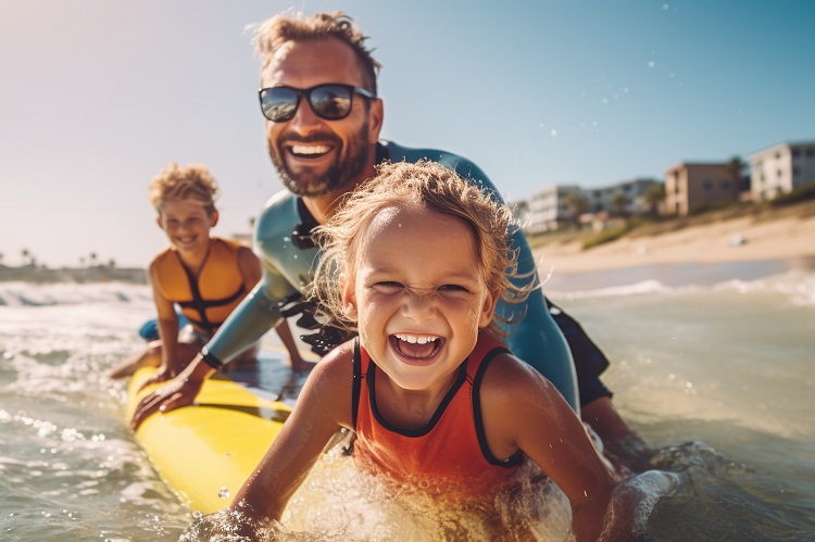 Vater, Tochter und Sohn mit Schwimmwesten auf Surfboard am Strand lachen in die Kamera.