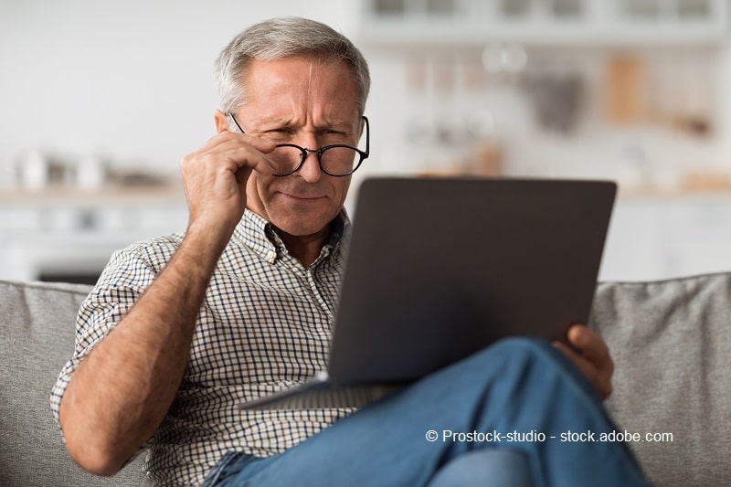 Bild eines älteren Mannes, der auf der Couch sitzt und angestrengt auf einen Laptop-Bildschirm sieht.