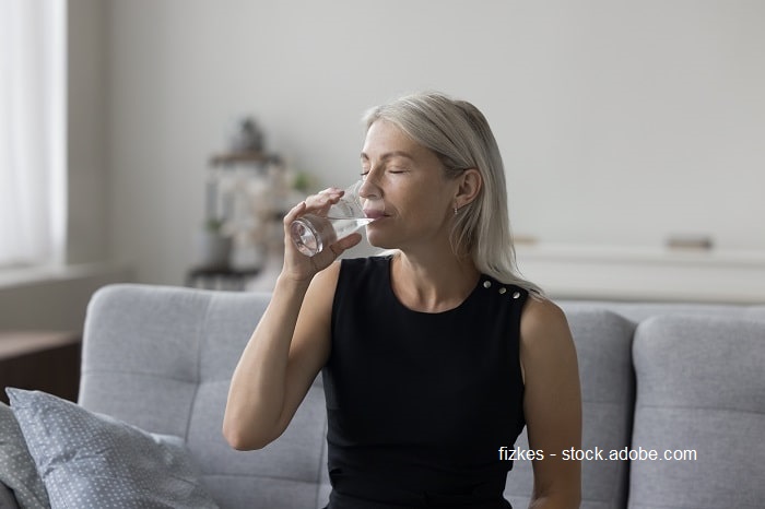 Bild einer Frau mit grauen Haaren, die auf der Couch sitzt und ein Glas Wasser trinkt.